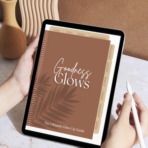 Könnte beinhalten: Ein digitales Tablet, das ein braunes Journal mit dem Titel "Goodness Glows" und dem Untertitel "The Ultimate Glow Up Guide" anzeigt. Das Journal hat Registerkarten für Quiz, Ziele, Budget und Manifestationen. Ein weißer Stift wird in der Nähe des Bildschirms gehalten.