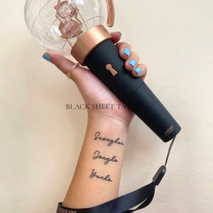 May include: A person's wrist with a temporary tattoo of the names "Seunghwan", "Jongho", and "Yunho" in cursive script. The person is holding a black and gold light stick with a globe on top.