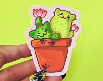 May include: A die-cut sticker featuring two green cat-shaped cacti sitting in a terracotta-colored pot. The cats have small pink flowers on their heads and bodies. The pot is decorated with dark brown paw prints and has a light pink border outline.