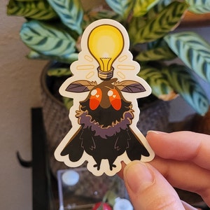 May include: A die-cut sticker featuring a cartoon moth with a lightbulb above its head. The moth is black with a purple collar, brown wings, and large orange eyes. The lightbulb is yellow with a white glow.