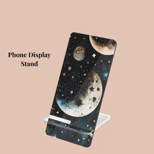 May include: A phone display stand with a celestial design. The stand features a black background with moons, stars, and a watercolor effect. The text "Phone Display Stand" is printed on the left side.