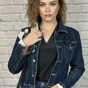 May include: A dark wash denim jacket with a tie-dye design on the collar and cuffs. The jacket has a classic design with buttoned pockets and a button closure. The model is wearing a black top and blue jeans.