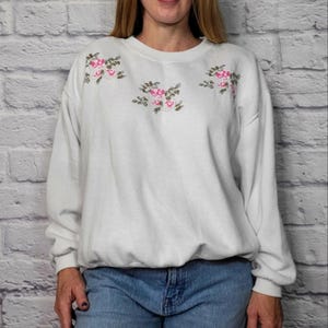 May include: White long-sleeved sweatshirt with embroidered pink and green floral designs. The crew neck sweatshirt has a ribbed hem and cuffs. The floral embroidery is on the chest and shoulders.