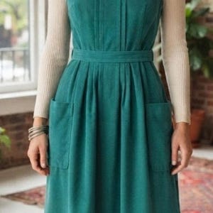 May include: A teal sleeveless corduroy pinafore dress with a square neckline, a fitted waist, and two side pockets, worn over a beige long-sleeved ribbed turtleneck sweater, paired with brown leather lace-up ankle boots and silver hoop earrings.