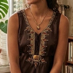May include: A sleeveless dark brown top featuring a V-neckline with intricate gold and copper-toned beadwork and embroidery. The garment includes a central decorative medallion detail at the chest and a relaxed fit, paired with blue denim jeans.