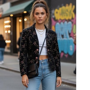 May include: A black blazer with a floral pattern and pink accents. The blazer has a classic lapel and pink buttons. The outfit includes a white top, light blue jeans, and a black crossbody bag. The model is wearing gold jewelry.