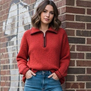 May include: A red knit sweater with a black zipper and collar trim. The sweater has long sleeves and is paired with blue jeans. The model is leaning against a brick wall.
