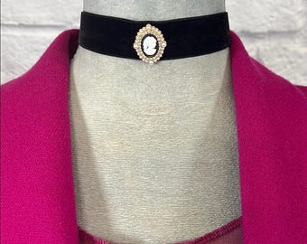 Op de afbeelding: Een zwarte fluwelen choker ketting met een camee hanger. De hanger heeft een wit profiel tegen een zwarte achtergrond, omringd door kleine witte kralen en een goudkleurig frame. De ketting wordt op een mannequin getoond.