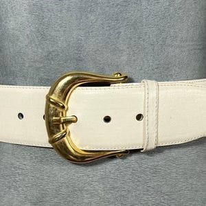 May include: Cream-colored leather belt with a large, gold-tone buckle. The buckle has a curved design. The belt has several holes for adjustment and a matching leather loop to secure the belt end. A classic fashion accessory.