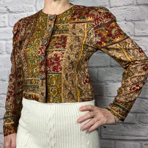 May include: A vintage patterned jacket with a button-up front. The jacket features a mix of red, gold, and brown colors with a floral and paisley design. The sleeves are long and the jacket is paired with a white skirt.