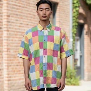 May include: A short-sleeved button-down shirt with a patchwork design. The shirt features a pattern of colorful squares in shades of pink, green, yellow, blue, and orange. The collar is yellow, and the shirt is worn by a person.