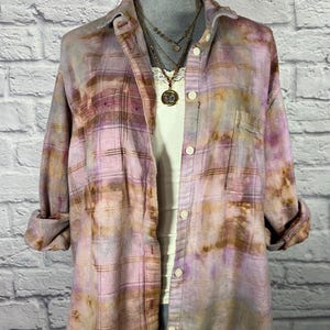 May include: A long-sleeve button-down shirt featuring a plaid pattern with tie-dye effects in shades of muted purple, brown, and cream. The shirt has a collar, a single chest pocket, and rolled cuffs, worn over a cream-colored tank top with lace trim.