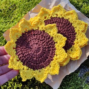 May include: Two crocheted sunflower coasters in brown and yellow. The coasters are made with a textured yarn and have a raised center.