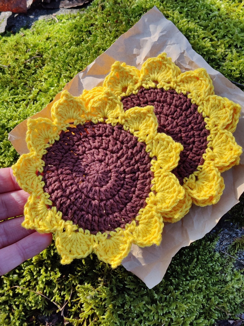 May include: Two crocheted sunflower coasters in brown and yellow. The coasters are made with a textured yarn and have a raised center.