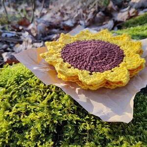 May include: A crocheted sunflower coaster in yellow and brown. The coaster is sitting on a piece of brown paper on top of green moss.