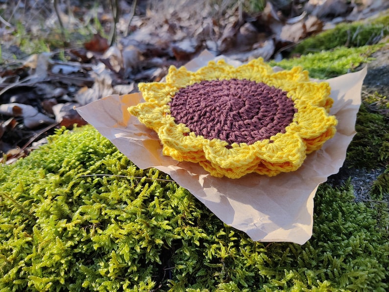 May include: A crocheted sunflower coaster in yellow and brown. The coaster is sitting on a piece of brown paper on top of green moss.