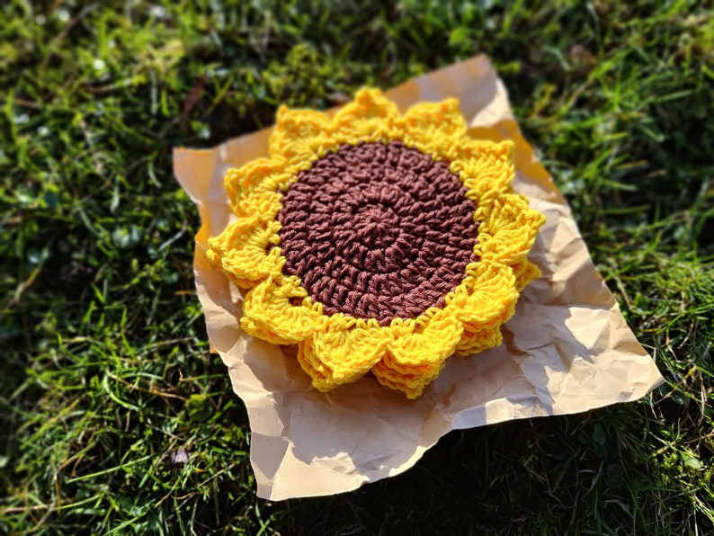 May include: A crocheted sunflower coaster in yellow and brown. The coaster is on a brown paper background.