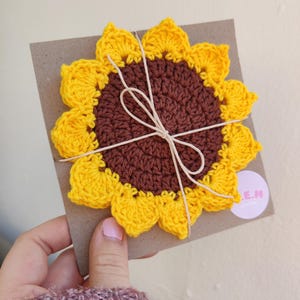 May include: A crocheted sunflower coaster with a brown center and yellow petals. The coaster is tied with twine and has a small white label with the text "E.M. Creations" on it.