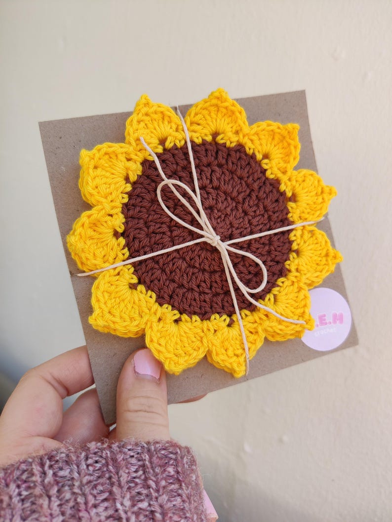 May include: A crocheted sunflower coaster with a brown center and yellow petals. The coaster is tied with twine and has a small white label with the text "E.M. Creations" on it.