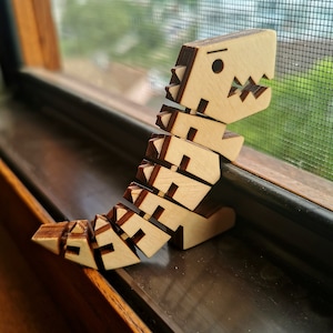 Puede incluir: Un juguete de dinosaurio de madera con un cuerpo segmentado, de pie en un alféizar de ventana. El dinosaurio es de color marrón claro y tiene una cara simple con un ojo negro.