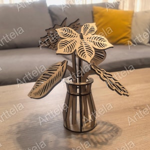 Può includere: Una composizione floreale decorativa in legno in un vaso. La composizione presenta foglie e fiori tagliati al laser in colore legno naturale. Il vaso è anch'esso in legno e ha una forma cilindrica. La composizione è su un tavolo di legno.