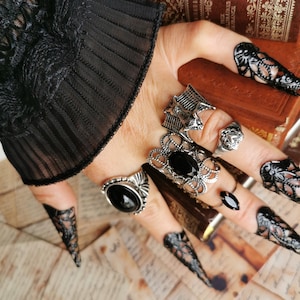 May include: A collection of silver-toned rings featuring black stones, a bat motif, and intricate filigree designs. The rings are worn alongside black lace fingerless gloves and decorative black filigree nail covers on each fingertip.