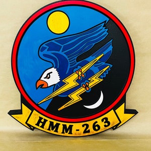 May include: A round wooden sign with a blue and black eagle design. The eagle is flying with lightning bolts in its talons. The sign has a red and yellow border and the text "HMM-263" in yellow letters.
