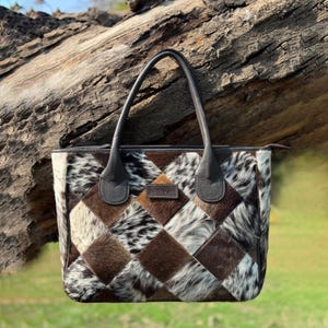 Op de afbeelding: Een patchwork tote bag met een geometrisch patroon van bruine, witte en zwarte vierkanten van koeienhuid. De tas heeft donkerbruine leren handvatten en afwerking, met een klein rechthoekig label aan de voorkant.