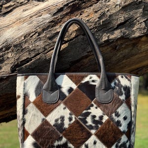 Op de afbeelding: Een patchwork tote bag met een geometrisch patroon van bruine, witte en zwarte koeienhuid vierkanten. De tas heeft donkerbruine leren handvatten en afwerking. De tas wordt buiten getoond.