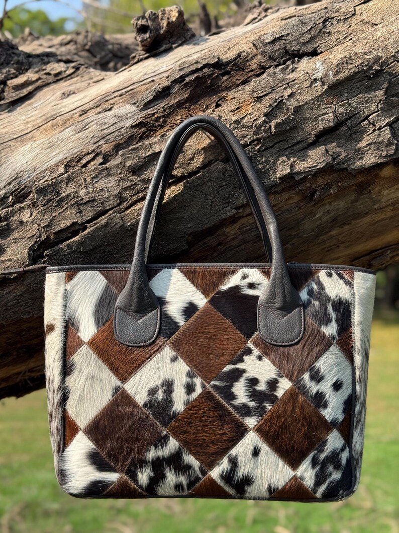 Op de afbeelding: Een patchwork tote bag met een geometrisch patroon van bruine, witte en zwarte koeienhuid vierkanten. De tas heeft donkerbruine leren handvatten en afwerking. De tas wordt buiten getoond.