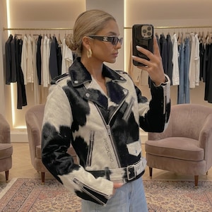 May include: A black and white cowhide print motorcycle jacket with silver zippers and a belt. The jacket has a classic biker style with a notched collar and asymmetrical zipper closure. The text "Chic Harbor" is printed on the jacket.