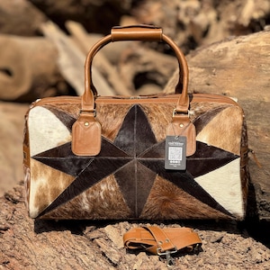 May include: A brown and tan leather duffel bag with a star design on the front. The bag has a top handle and a detachable shoulder strap. The bag is made of cowhide and has a zipper closure. A tag with text is attached to the bag.