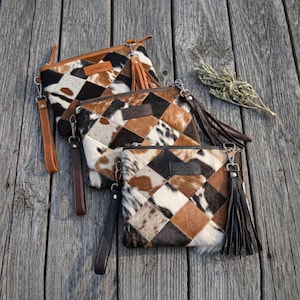 Könnte beinhalten: Drei Clutch-Taschen aus Kuhfell mit einem Patchwork-Design in Braun-, Schwarz- und Weißtönen. Jede Tasche hat eine Handschlaufe und einen Quastenanhänger. Die Taschen sind auf einer verwitterten Holzoberfläche angeordnet.