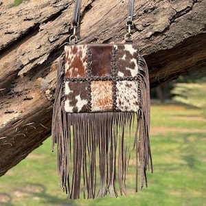 May include: A brown leather crossbody bag with a patchwork design. The bag features a mix of brown and white cowhide squares, stitched together. Long brown fringe hangs from the bottom. The bag has a brown leather strap and gold-coloured hardware.