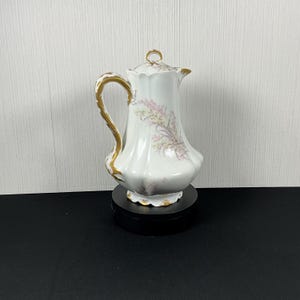 May include: White porcelain coffee pot with a gold handle and accents. The pot features a floral design in pink and yellow. The lid is also white with a gold handle. The pot is sitting on a black base.