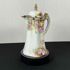 May include: A white porcelain coffee pot with a lid and handle, adorned with pink and yellow rose floral patterns. The pot has gold accents on the handle, lid, and rim. It is displayed on a black circular base.