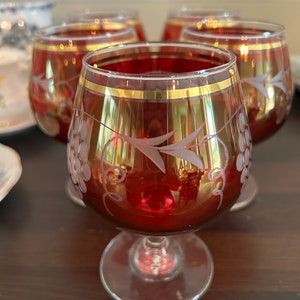 Può includere: Set di quattro bicchieri da brandy con bordo rosso e oro. Ogni bicchiere presenta un gambo trasparente e un motivo a uva inciso. I bicchieri sono disposti su una superficie di legno scuro, con altre stoviglie sullo sfondo.