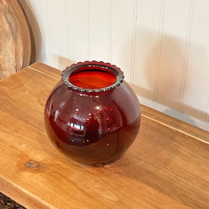 Peut inclure: Un vase rond en verre rouge avec un bord festonné. Le vase a une finition brillante et une couleur rouge foncé. L'intérieur du vase est orange vif. Le vase est posé sur une surface en bois.