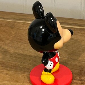 May include: A Mickey Mouse figurine, with a black head and ears, a tan face, and a red and white outfit. The figurine stands on a red circular base and has yellow shoes. The classic cartoon character is in a side profile.