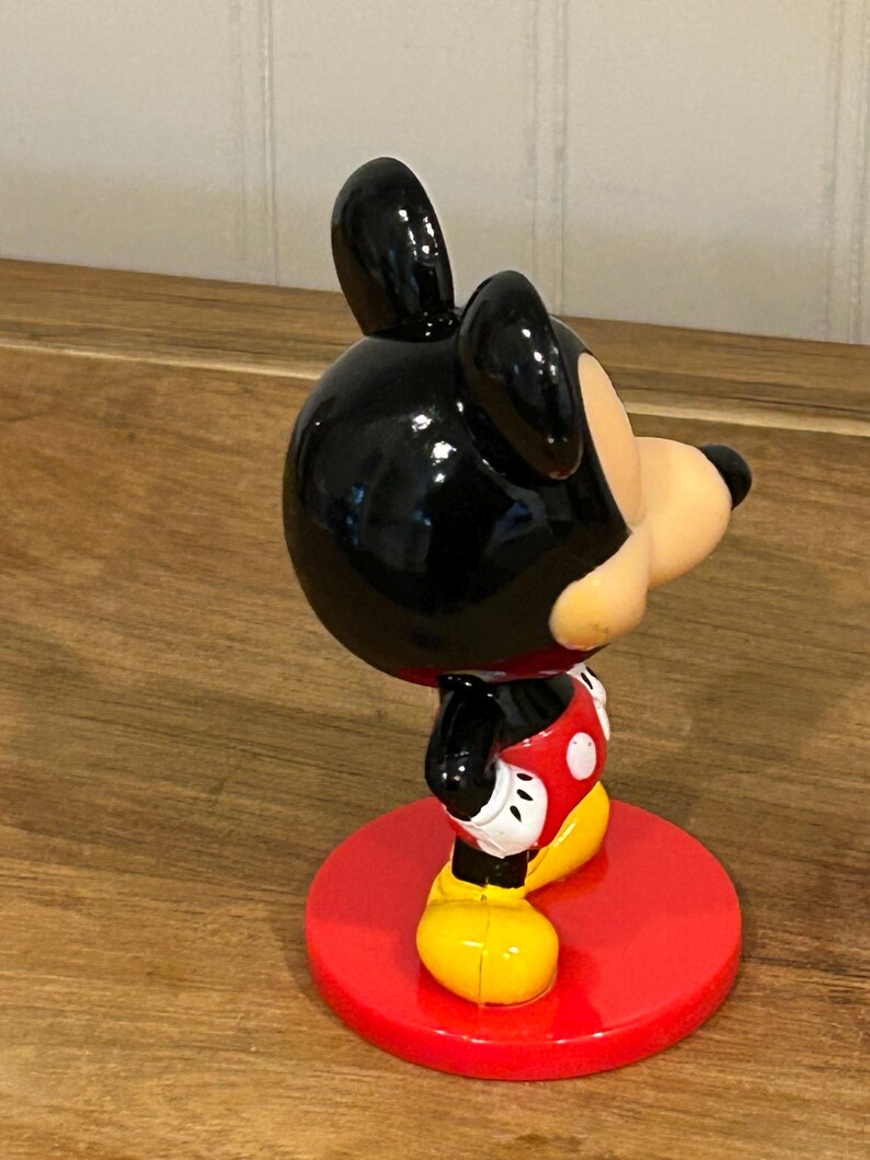 May include: A Mickey Mouse figurine, with a black head and ears, a tan face, and a red and white outfit. The figurine stands on a red circular base and has yellow shoes. The classic cartoon character is in a side profile.