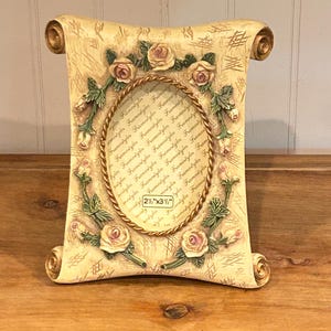 May include: A cream-colored scroll-shaped picture frame featuring a central oval opening with a rope-style border. The frame is decorated with sculpted pink roses and green leaves, and includes a label stating 2 1/2" x 3 1/2".