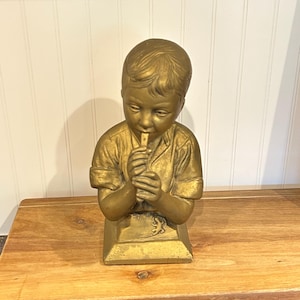 Op de afbeelding: Een goudkleurig beeld van een jonge persoon die fluit speelt. Het beeld staat op een rechthoekige basis en is gemaakt van een metalen materiaal. De figuur draagt een shirt met korte mouwen. Het beeld staat op een houten oppervlak.