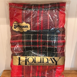 May include: A rectangular tablecloth in a red, black, and gold plaid pattern, packaged in clear plastic. The label reads New Softspun and Holiday Designs for Dining. The dimensions printed on the packaging are 60 inches by 104 inches.