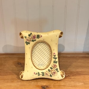 May include: A cream-colored scroll-shaped picture frame with gold-toned accents at the corners. The frame features raised floral motifs of pink and yellow roses. A central oval opening displays a paper insert with the text 2 1/2" x 3 1/2".