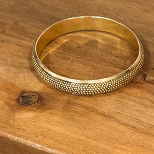 May include: A circular gold bangle bracelet featuring a textured, dotted surface pattern along the outer band. The interior of the bracelet is smooth and polished, reflecting a bright metallic gold finish throughout the entire piece of jewelry.