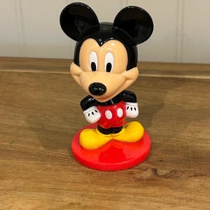 May include: A Mickey Mouse figurine with a black head and ears, a tan face, and a red and white outfit. The figurine stands on a red circular base with yellow shoes. The figure is a classic Disney character.