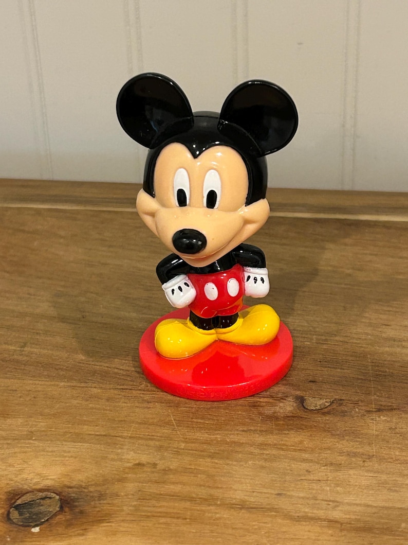 May include: A Mickey Mouse figurine with a black head and ears, a tan face, and a red and white outfit. The figurine stands on a red circular base with yellow shoes. The figure is a classic Disney character.