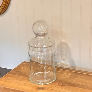 Peut inclure: Un bocal d'apothicaire en verre transparent avec un couvercle arrondi et un dessus bulbeux. Le bocal a une base large qui se rétrécit vers l'intérieur puis s'écarte à nouveau. Il repose sur un plateau en bois.