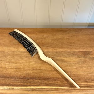 May include: A cream-colored hair comb with black bristles. The comb has a long, pointed handle and a curved head. The comb is made of plastic and is designed for styling hair. The comb is on a wooden surface.