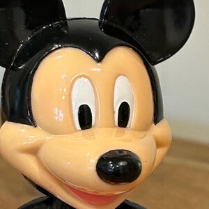 May include: A close-up of a Mickey Mouse figurine. The figurine has a black head and ears, a tan face, and a black nose. The figurine is wearing a black jacket with a red bow tie.